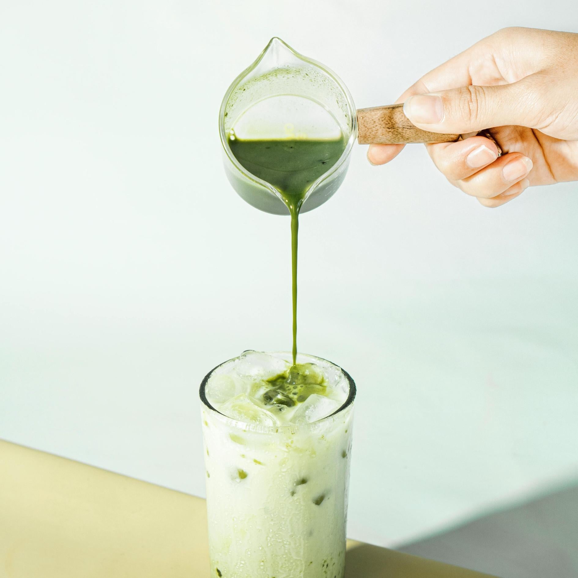 Hand pouring green liquid from a small pitcher into a glass with ice on a light background
