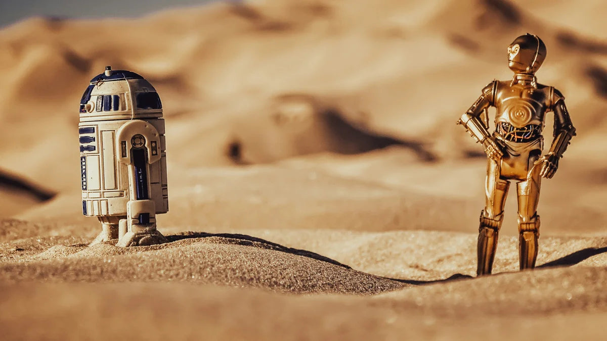 Two robot figures, R2-D2 and C3PO, standing on a sandy desert landscape.